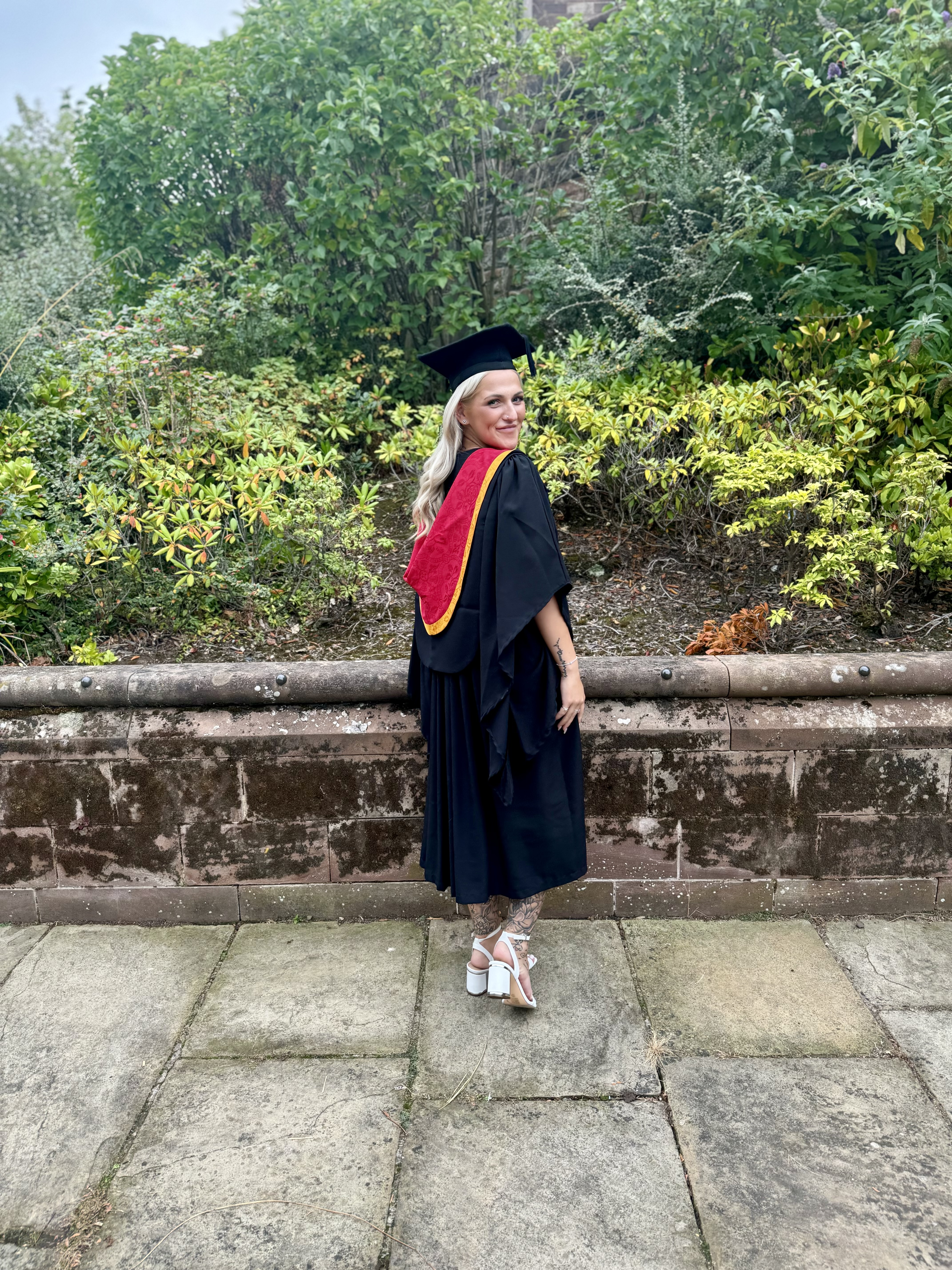 Graduate Kizzy standing outside with leaves in background wearing graduation cap and gown facing away from camera and looking back at camera and smiling