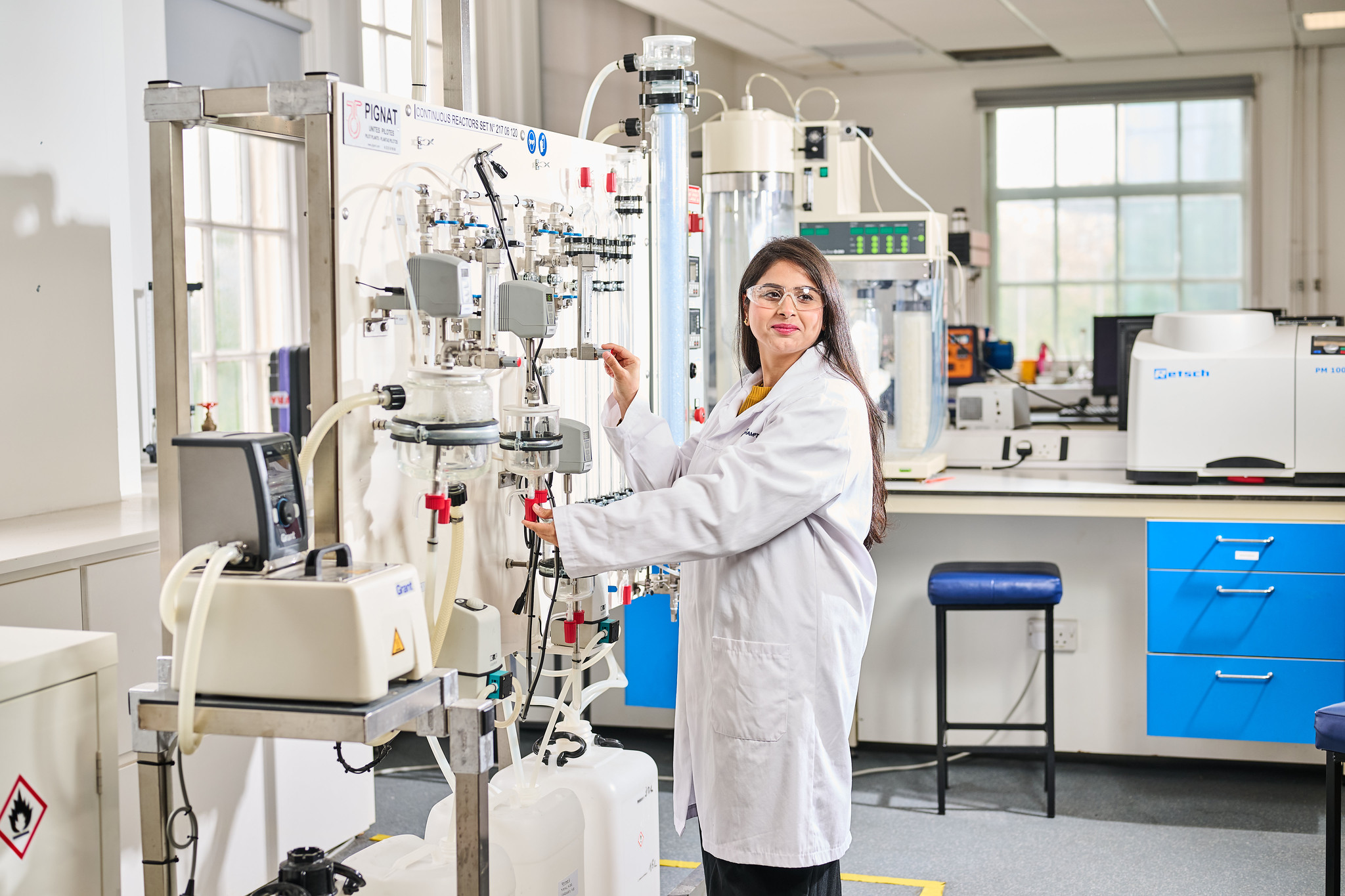 Student in a white lab coat standing in a chemical engineering lab at City Campus.