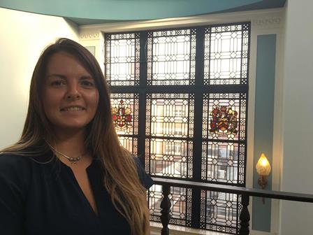 Clare Dickens photographed in front of a banister, a window behind her with two stained-glass coat of arms designs.