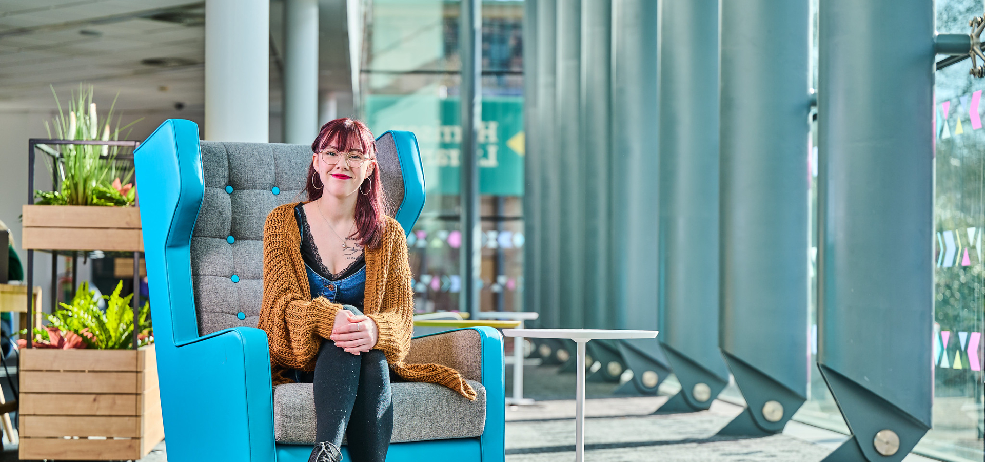 A student with pink hair wearing an orange cardigan, sitting in a blue armchair next to a small white table. Large windows are letting bright sunlight into the room, and an array of plants can be seen behind the student.