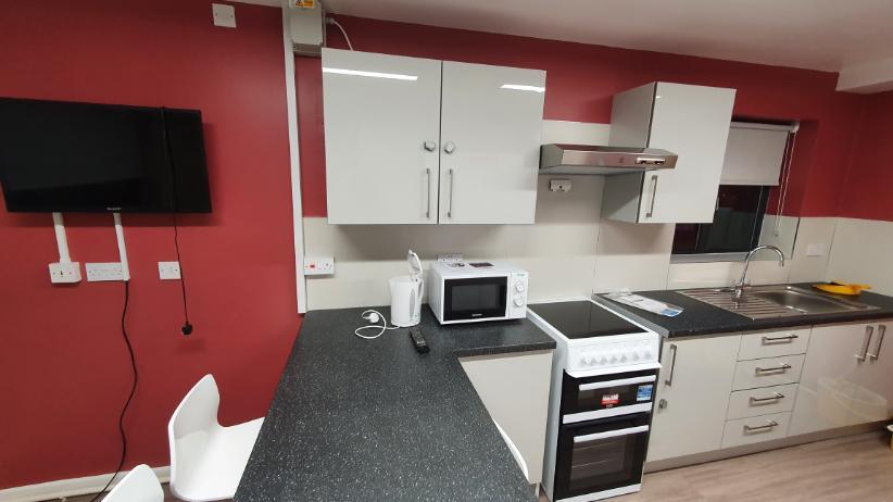 A kitchen in Telford Campus accommodation, with a TV on the wall and a countertop with a microwave and kettle on top of it, a sink, an oven with a hob, a sink and several cupboards and drawers