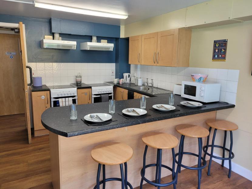 A kitchen in Walsall Campus accommodation, four chairs pulled up to a table attached to a countertop with a sink, microwave, kettle and hob on it, two ovens and several cupboards built into it