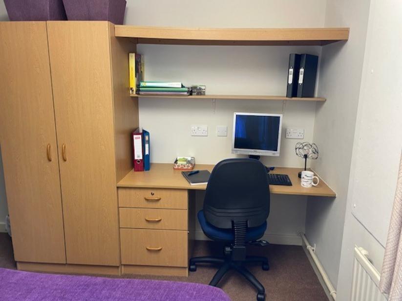 A desk in Telford Campus accommodation, with a chair pulled under it, shelving above it and a wardrobe and chest of drawers attached to it on the left
