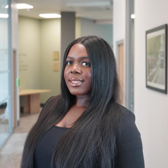 Photograph of Adedapo Fasinro standing in a corridor, wearing a black blouse.