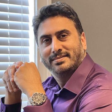 Jameel Ahmed wearing a purple shirt, posed over a table with a laptop.