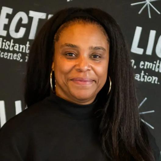 Photograph of Kerensa Hodge wearing a black jumper, standing in front of a blackboard lined with terminology related to lighting.