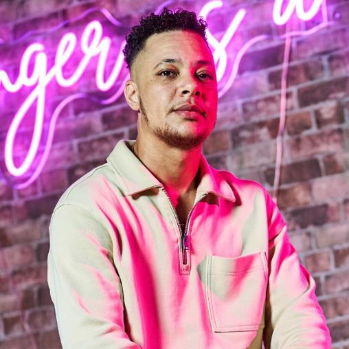 Nate Ethan Watson posed against a pink neon sign, wearing a white jacket.