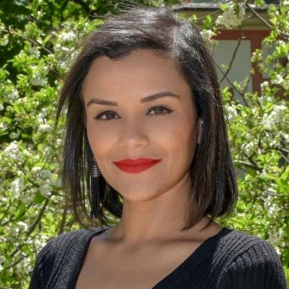 Nathalie Flores standing between two tall hedges, wearing a black blazer with a multicoloured brooch