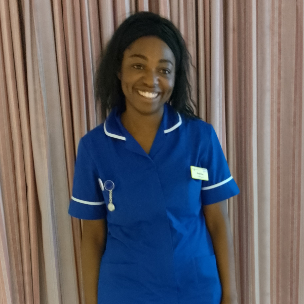 Sabrina Ffrench in a nursing uniform standing in front of a curtain.