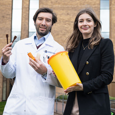 Profile photo of University of Wolverhampton alumni Olivia Simpson and Mattia Parati. Mattia is wearing a white lab coat and holding various 3D printed medical products. Olivia is wearing a black jacket with gold accents and is holding a yellow container with an orange lid.