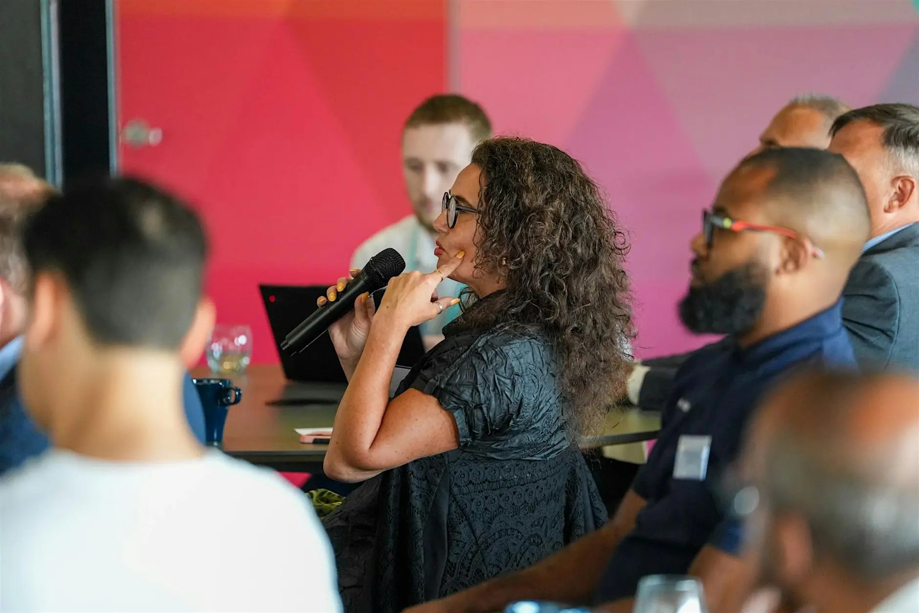 A woman with long, dark curly hair and glasses is seated at a business event. She is asking a question whilst holding a microphone