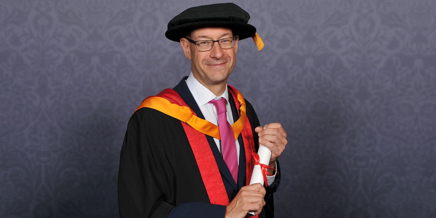 Honorary Graduate Neil Lloyd looking at the camera with his award