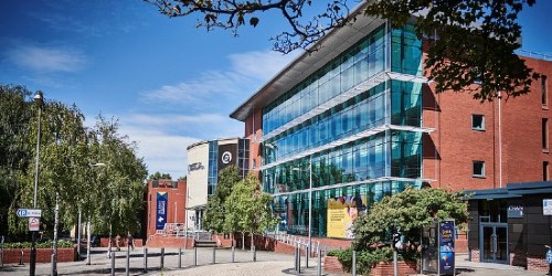 Exterior of the Ambika Paul building on a sunny day, a red-brick building with a large wall of windows currently decorated with a large graphic of a student.