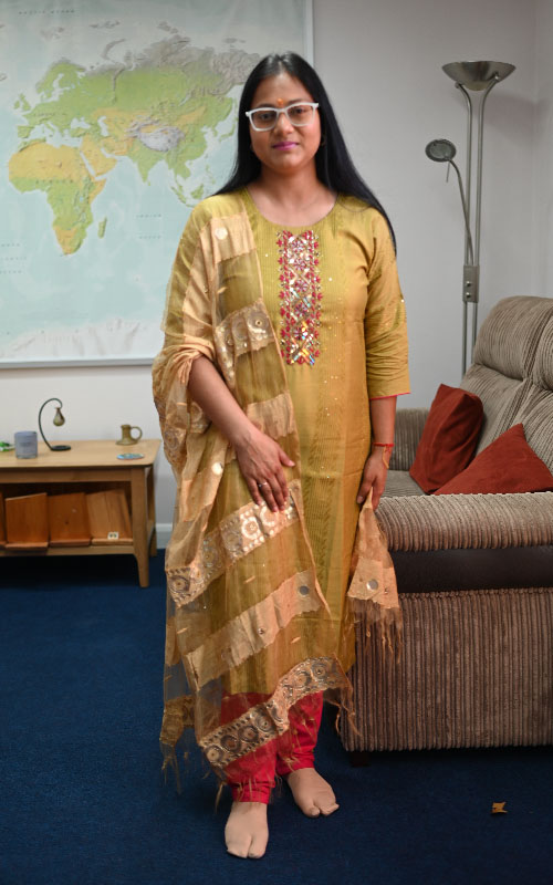 a full portrait photograph of student, Vanjyotsna Grover, wearing a yellow sari standing in the Chaplaincy in front of a world map