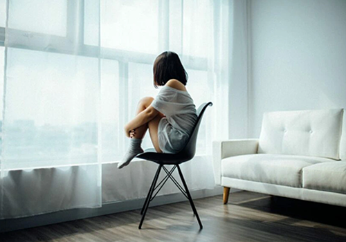 woman in white lounge wear perched on a chair looking longingly out of a window covered with white sheer curtain