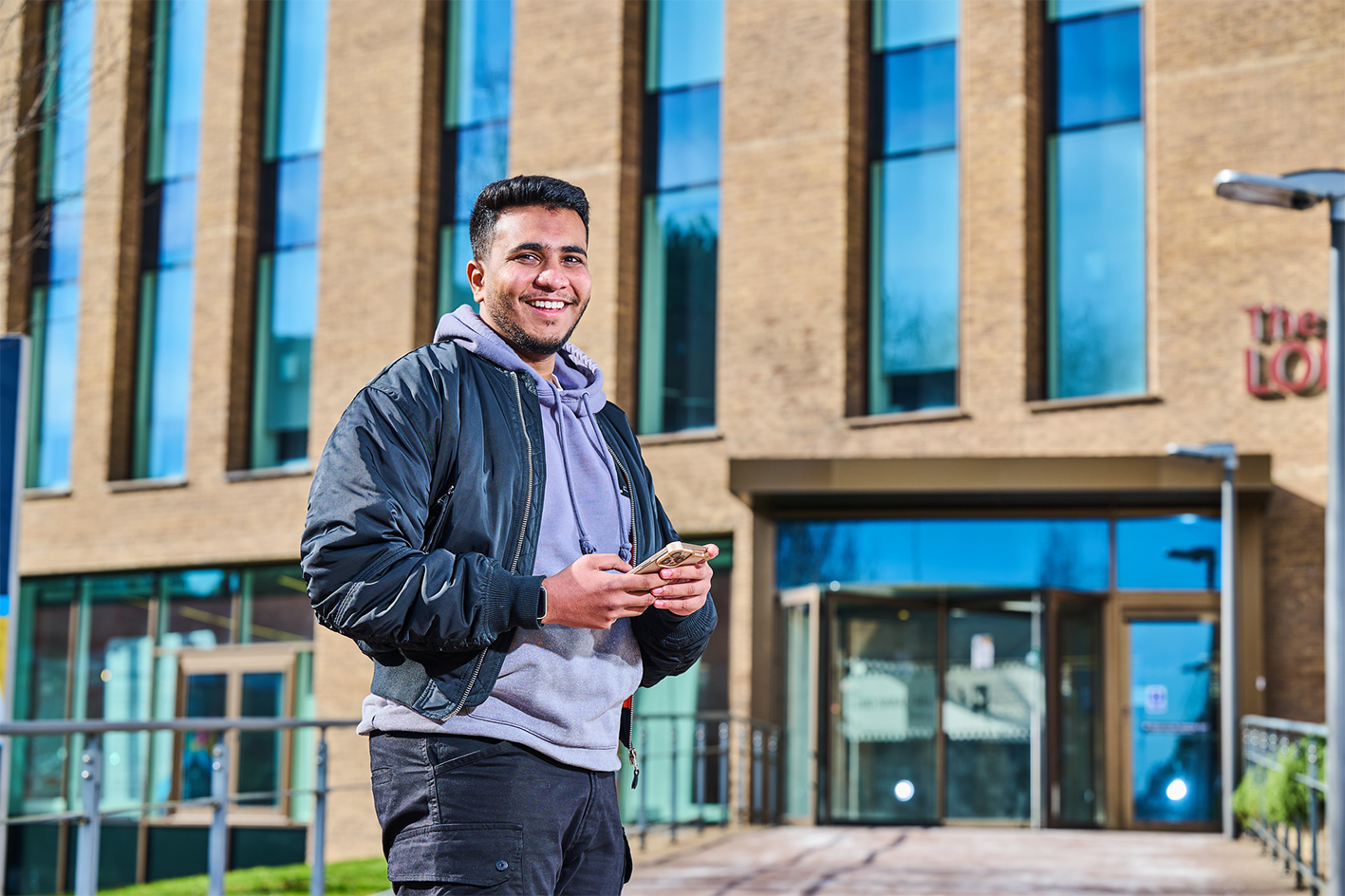 male student holding phone outside Lord Swraj Paul Building Business School