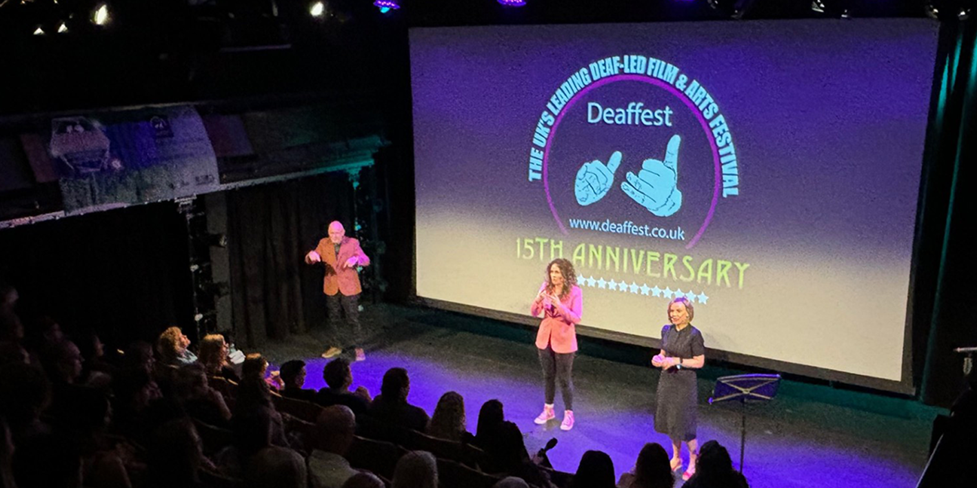 A photograph taken in a theatre at Deaffest. The photo is taken from the audience perspective looking onto a stage where three people are singing in BSL. The backdrop on the stage reads 