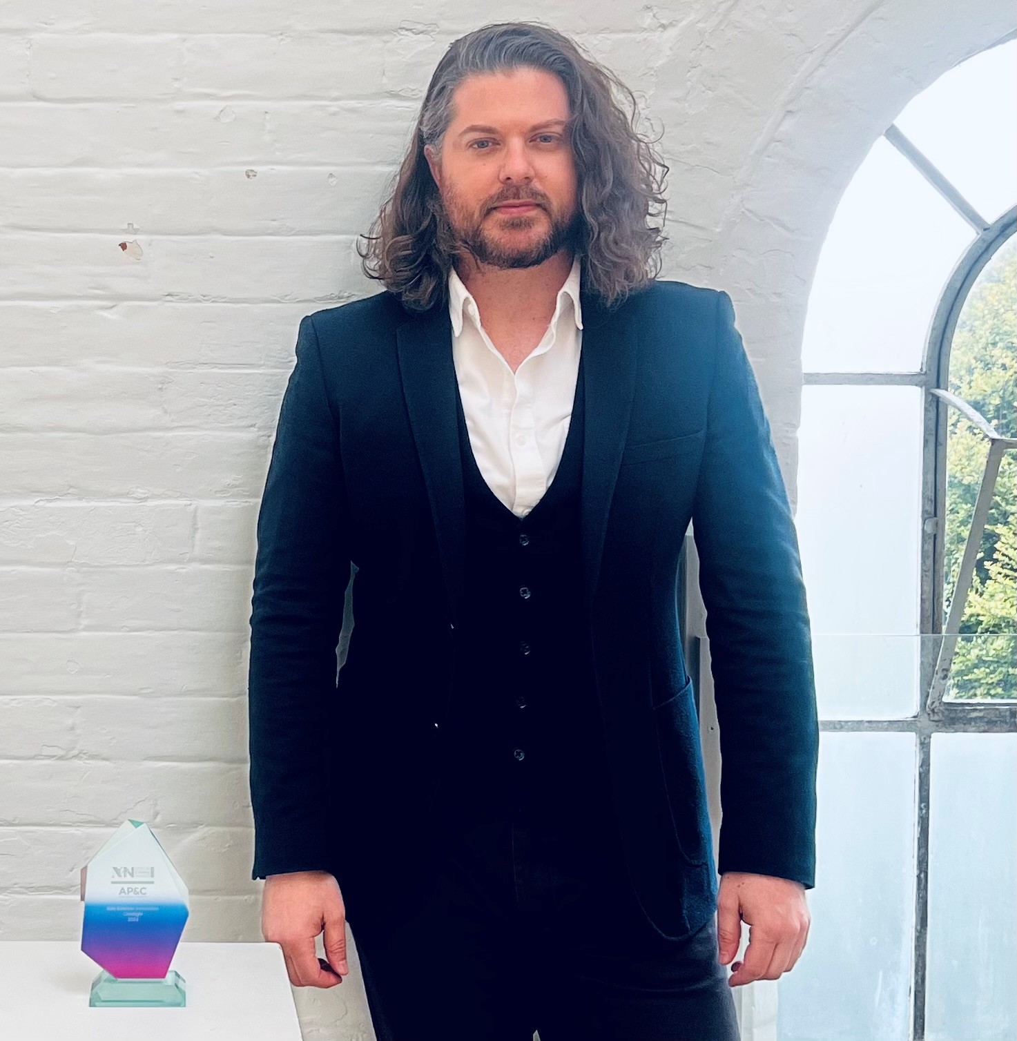 MsC psychology alum, Leon Mitchelle, dressed in a suit, stands against a white brick wall. An award is on the table to his side.