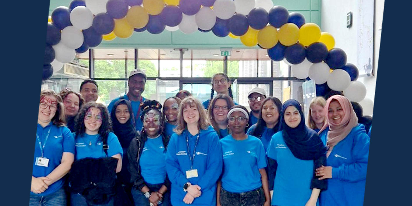 Photo of a group of smiling Student Ambassadors in blue tee-shirts under an arch of balloons