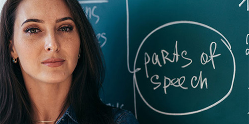 Woman standing in front of a blackboard with the words 'parts of speech' written in chalk
