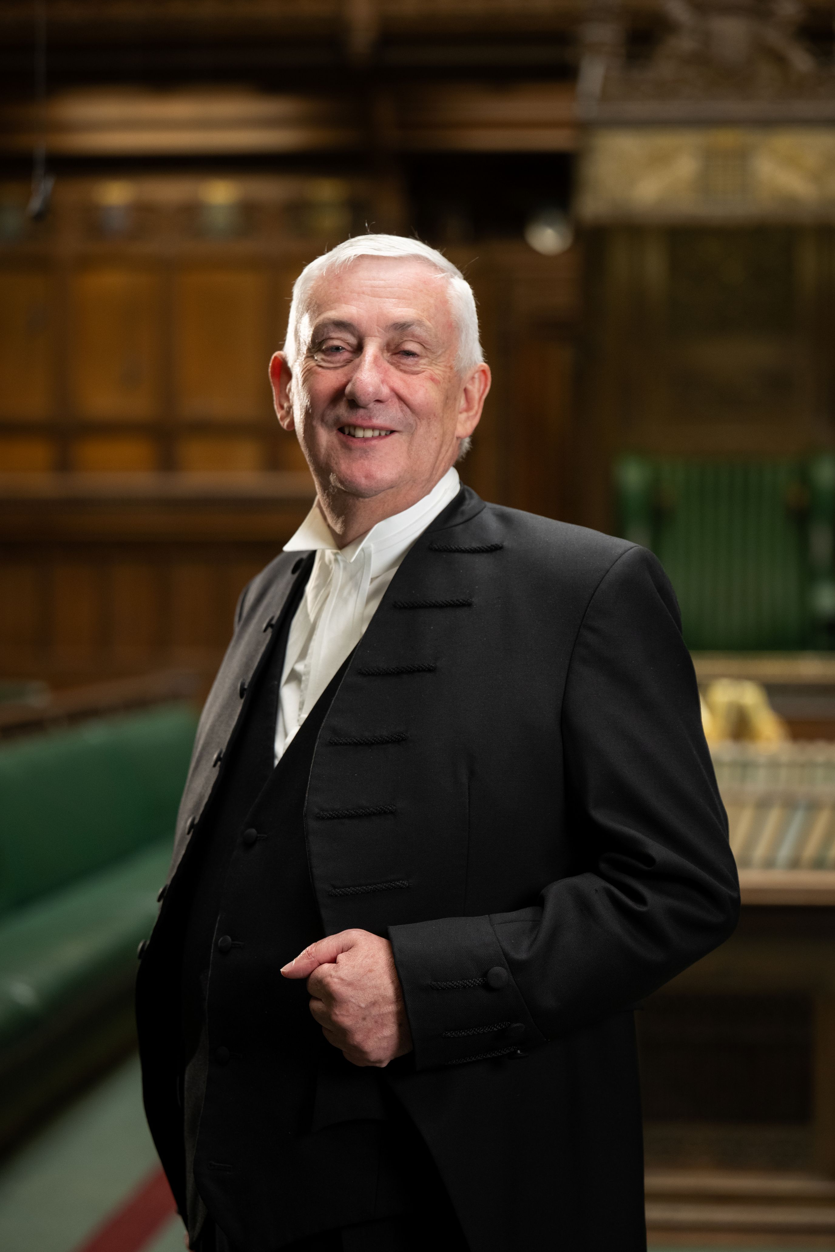 Sir Lindsay Hoyle is dressed in a dark suit and light shirt with the top botton undone. Behind him are the benches in the House of Commons