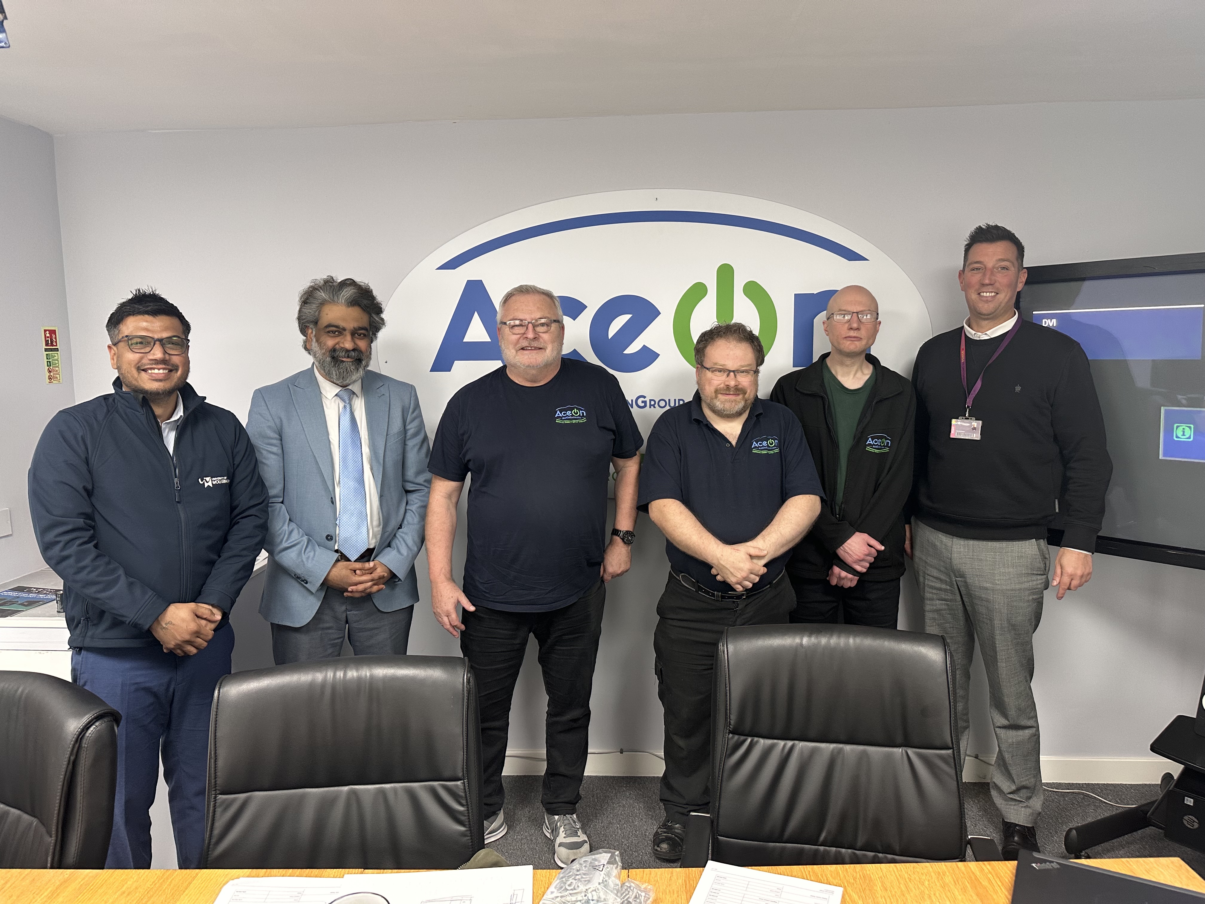 Representatives from Aceon and the University of Wolverhampton line up in front of the Aceon logo on the wall behind in green and blue lettering. The wear business casual clothing and smile for the camera