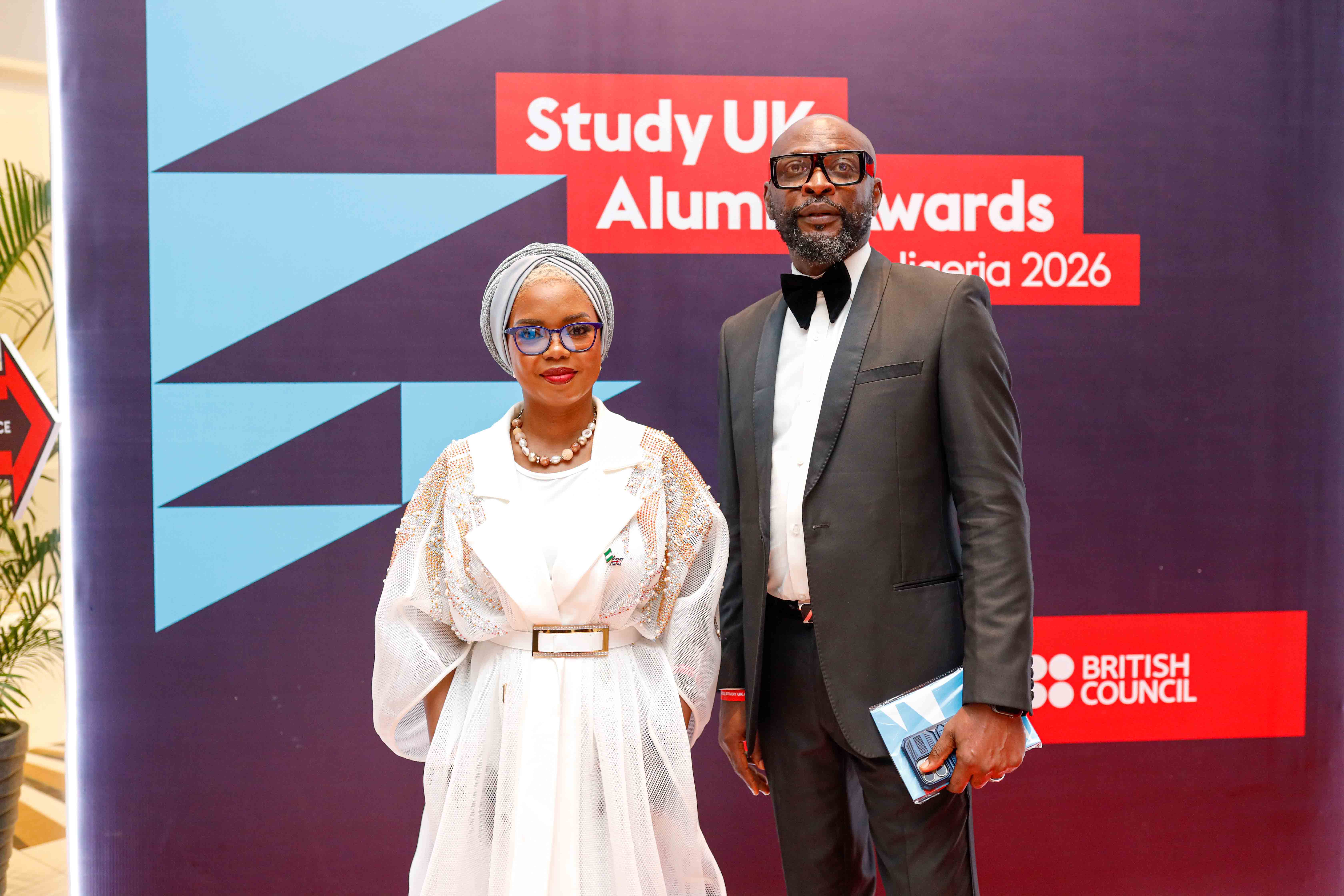 Sesi Oluwatoyin Orere in a white dress and grey headdress stands on the right of Chima Victor Kalu on the red carpet at the Alumni Awards in Nigeria