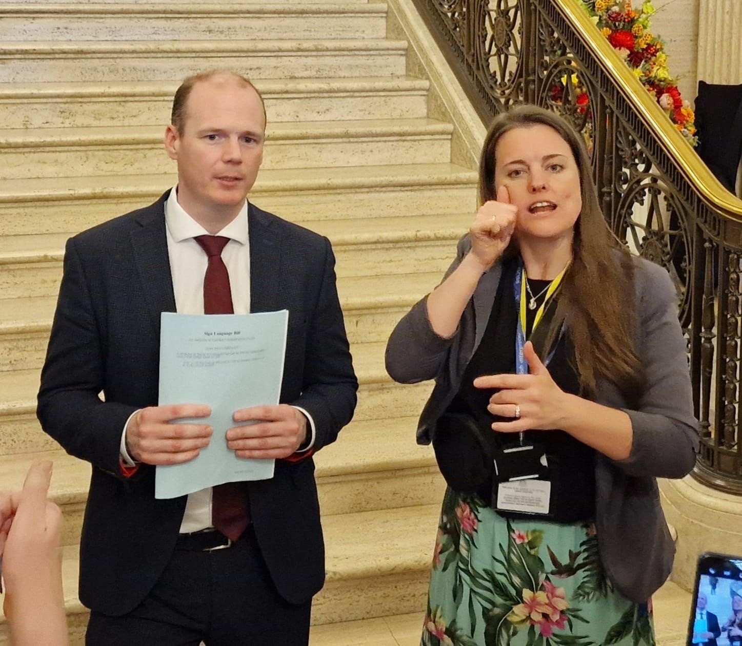 Gordon Lyons MLA stands in a suit in front of some steps, he is holding some paper and speaking to cameras. Sally Gillespie is beside him interpreting for the cameras.