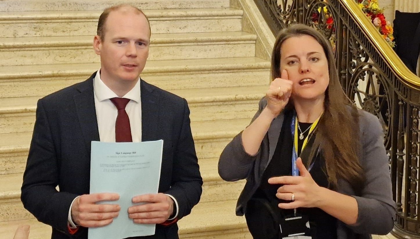 Gordon Lyons MLA stands in a suit in front of some steps, he is holding some paper and speaking to cameras. Sally Gillespie is beside him interpreting for the cameras.