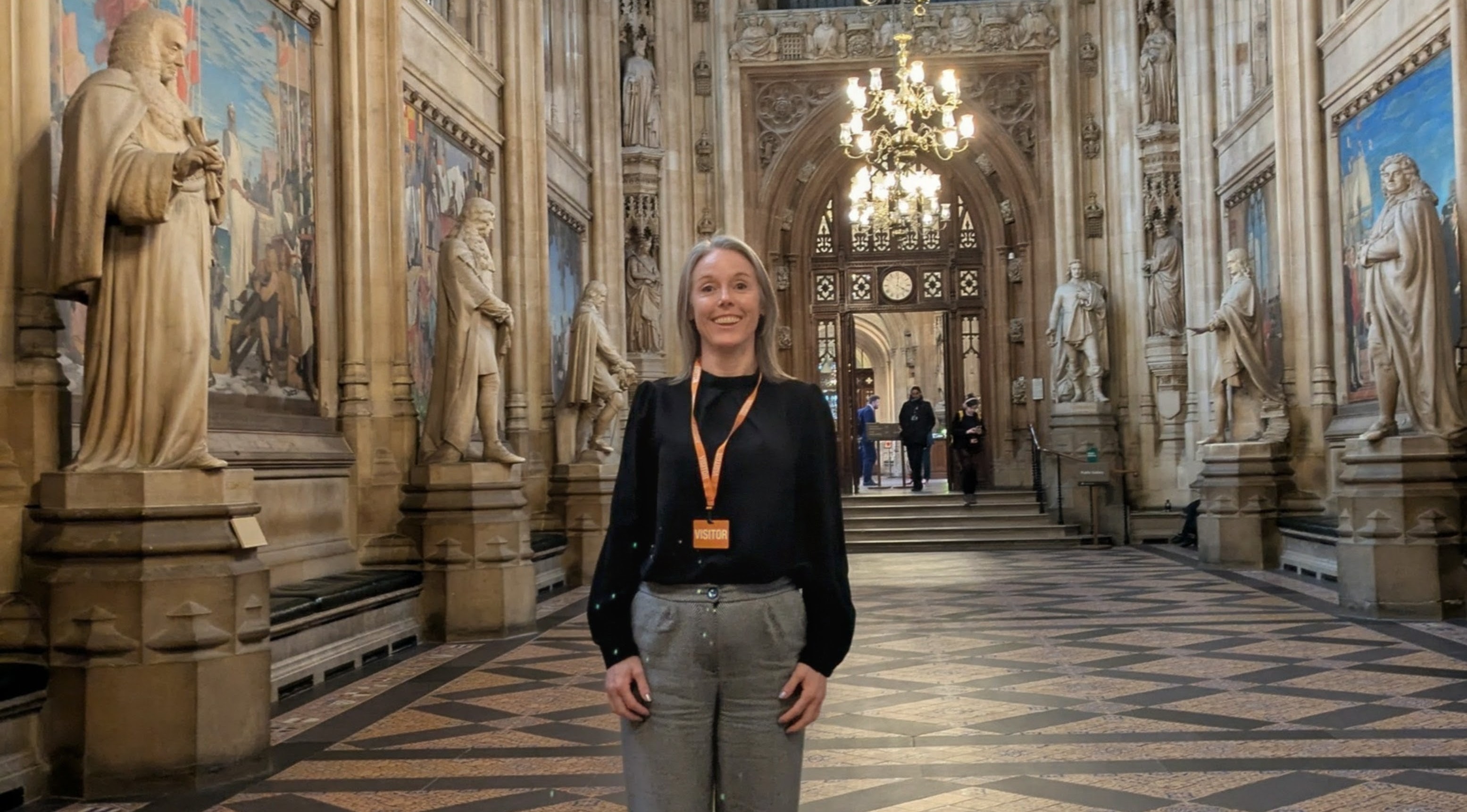 Becky stands in an ornate hall surrounded by marble statues and historic art works. There is a chandelier above her and the floor is covered in mosaic tiling.