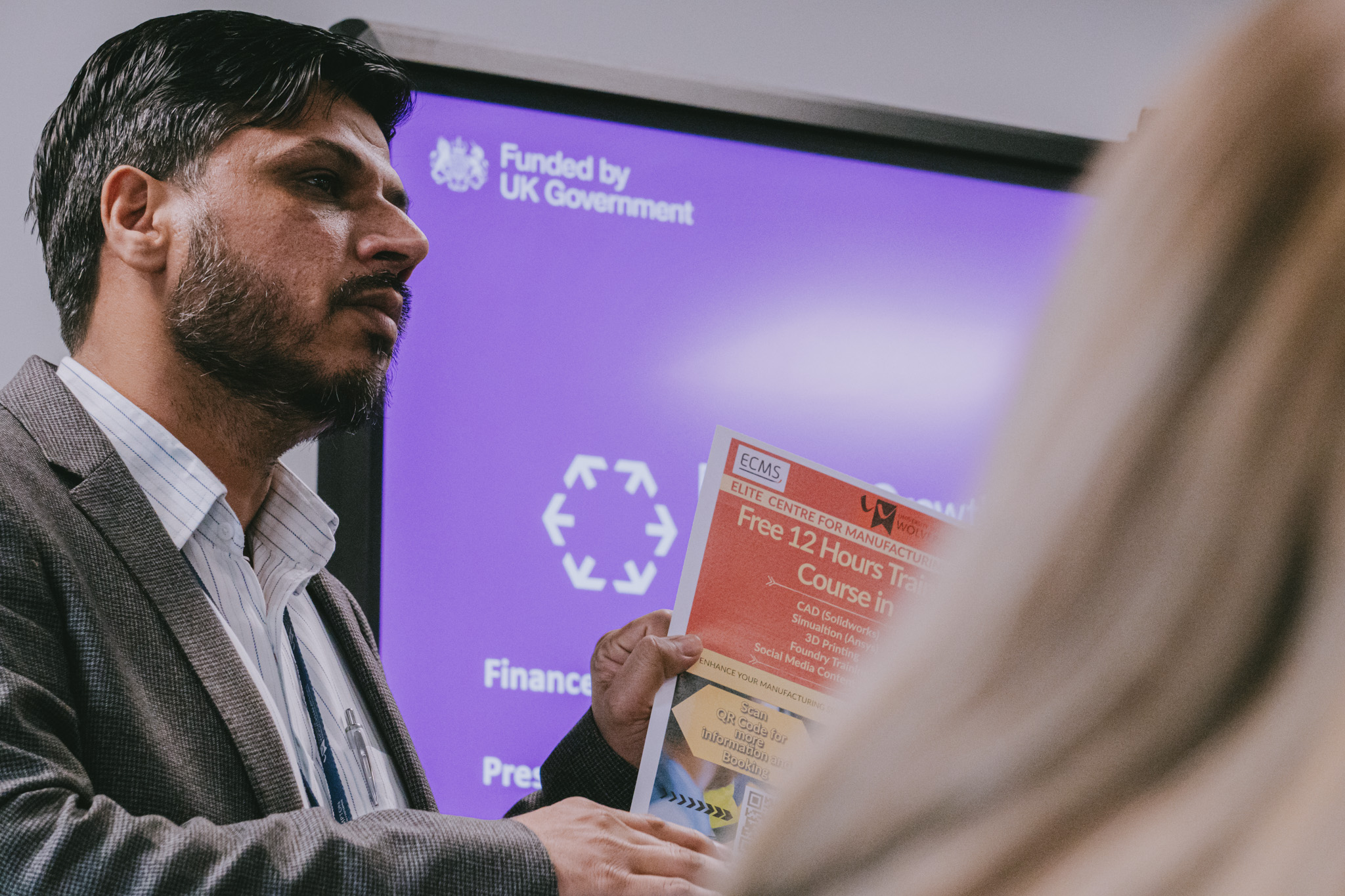 A man stands in front of a projector screen that display the Funded by UK Government logo. He is holding a brochure whilst speaking to a delegate at a business event