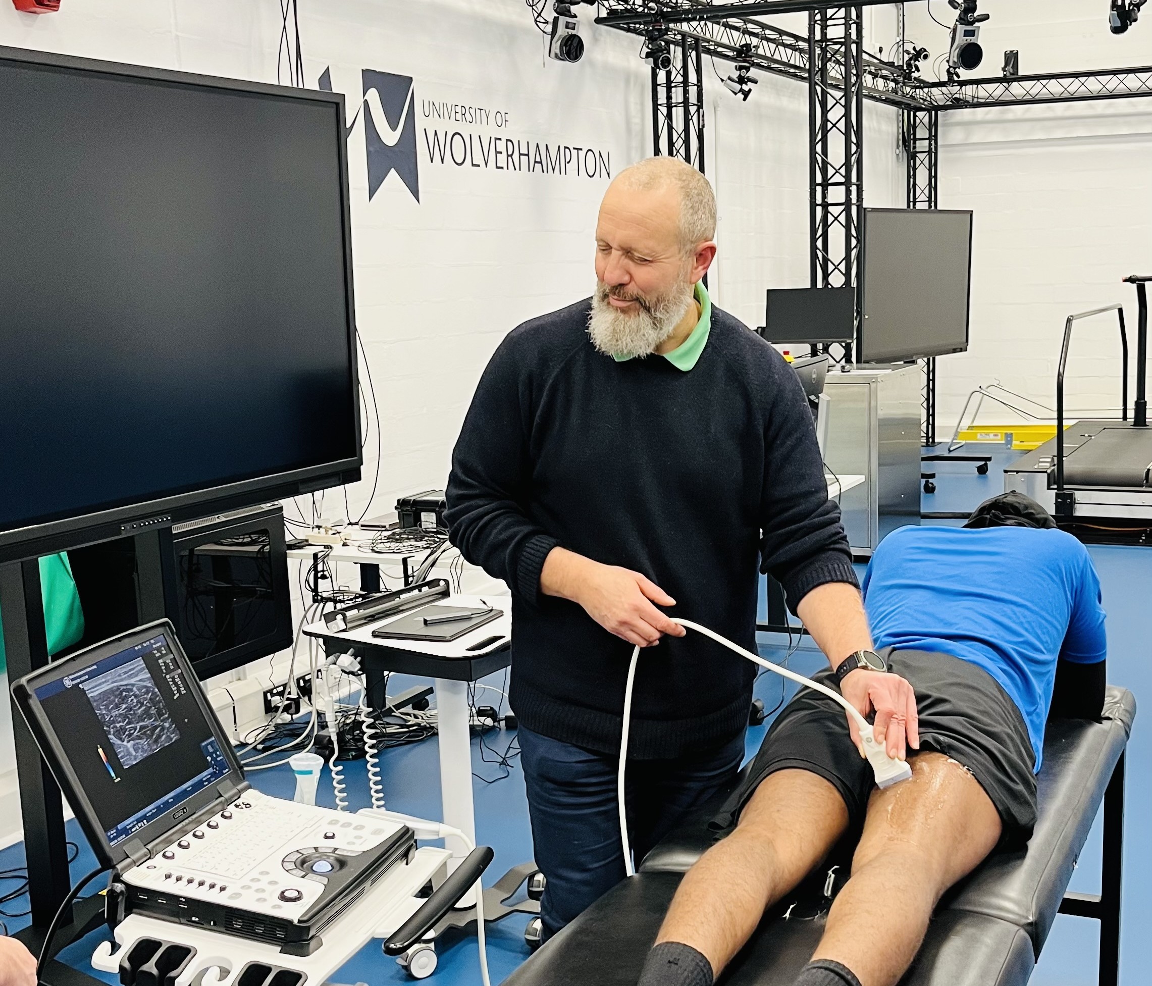 Professor Matthew Wyon, Professor of Exercise Physiology stands next to a therapy table where a male lays facedown. Matthews is using a ultrasound scan on the rear calf of the male and looking at a computer screen.