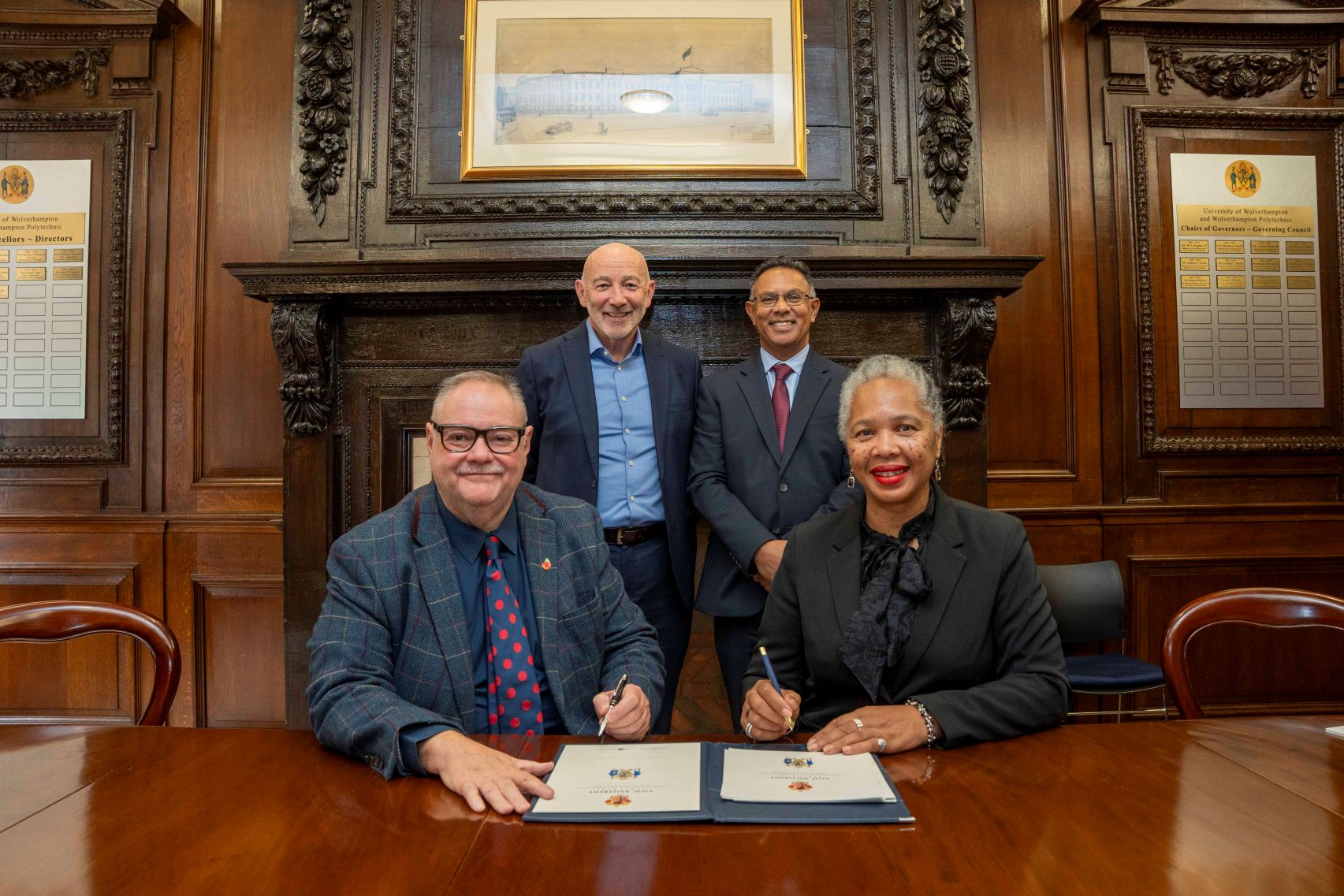 Representatives from the council and university are in an old panelled room. Two are seated with two standing behind them. On the table in front is the agreement they are signing.