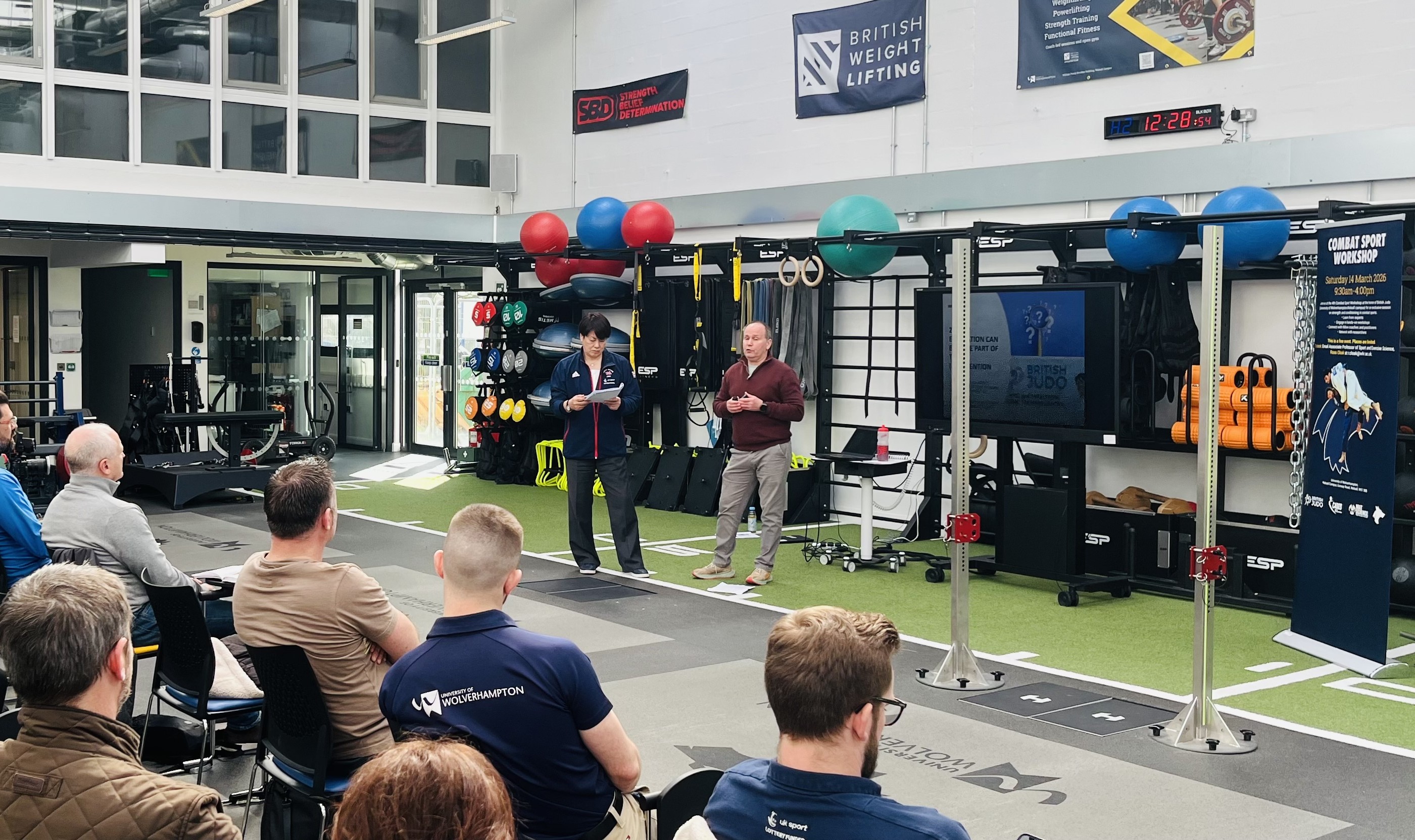 A group of people sit facing a presentation taking place inside a spacious indoor sports performance facility. Two presenters stand at the front on a turf surface, with one holding a tablet and the other speaking. Behind them is an array of training equipment, including kettlebells, medicine balls, weight plates, resistance bands, and exercise machines