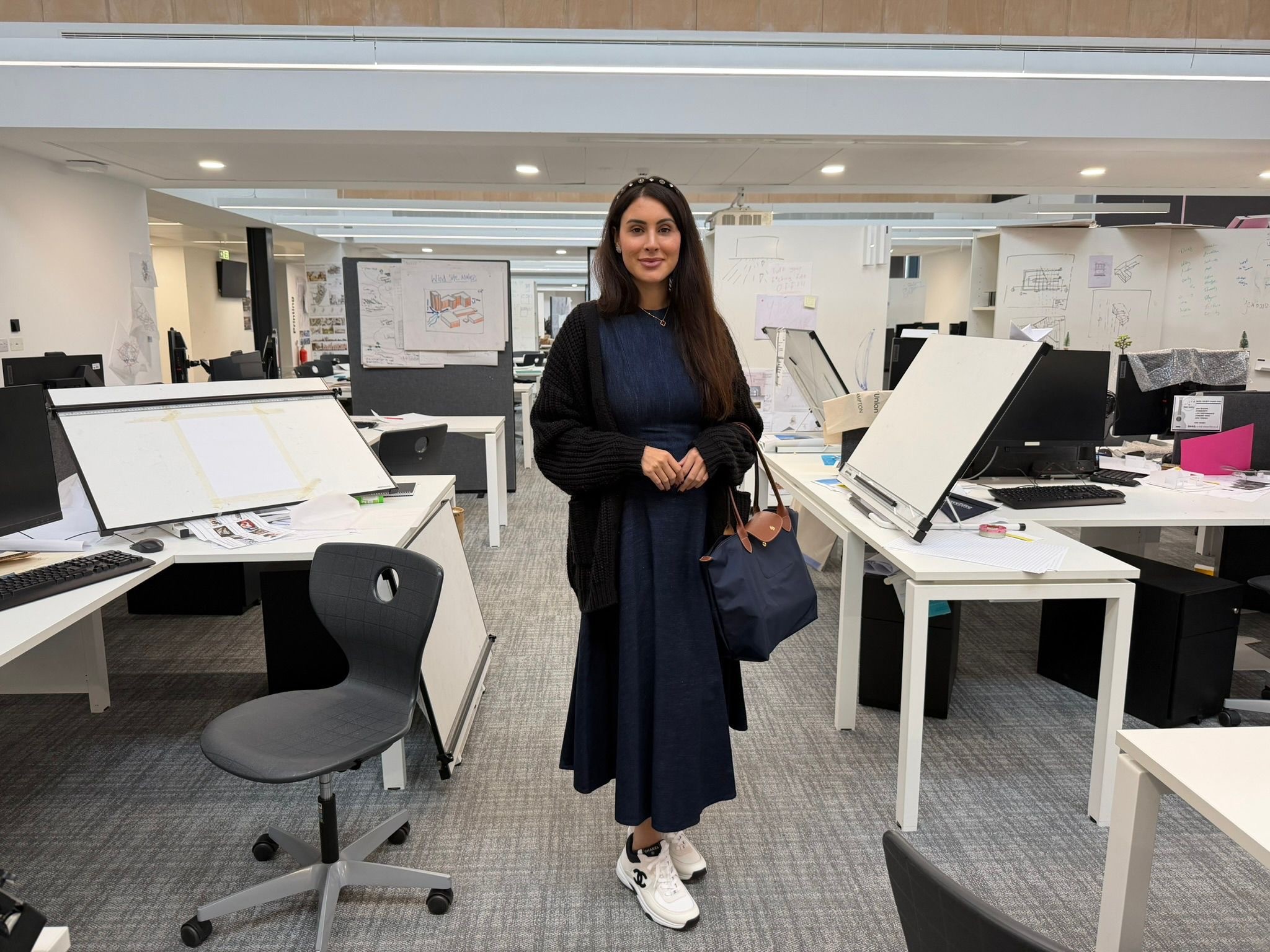 Deanna stands in an office setting, she has desks either side of her. She's wearing a long blue dress, white trainers and an Alice band in her hair.