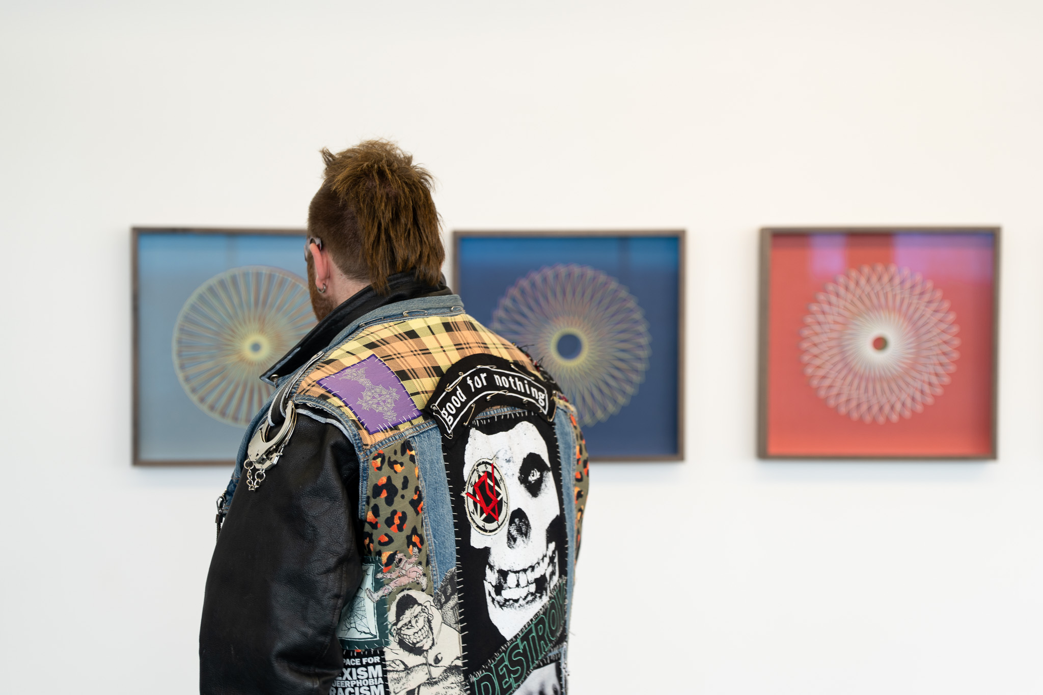 Back view of a man with a grown out Mohican hairstyle who wears a leather jacket with skull motif on colourful patchwork on the back. He look at three pieces of artwork on a wall in front of him