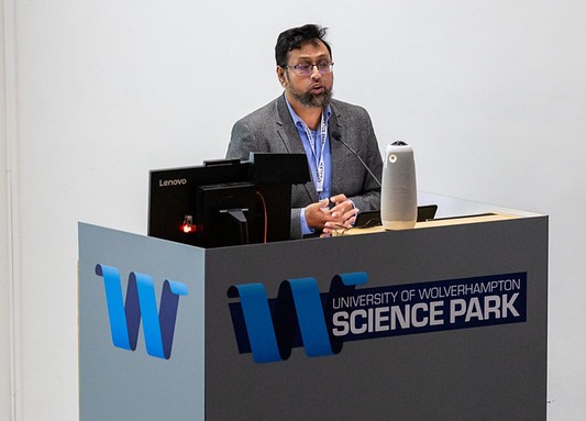 Dr Rahman, research project lead stands behind a grey podium with the University of Wolverhampton Science Park logo on the front. He is addressing an audience and wears a grey suit with a blue shirt.