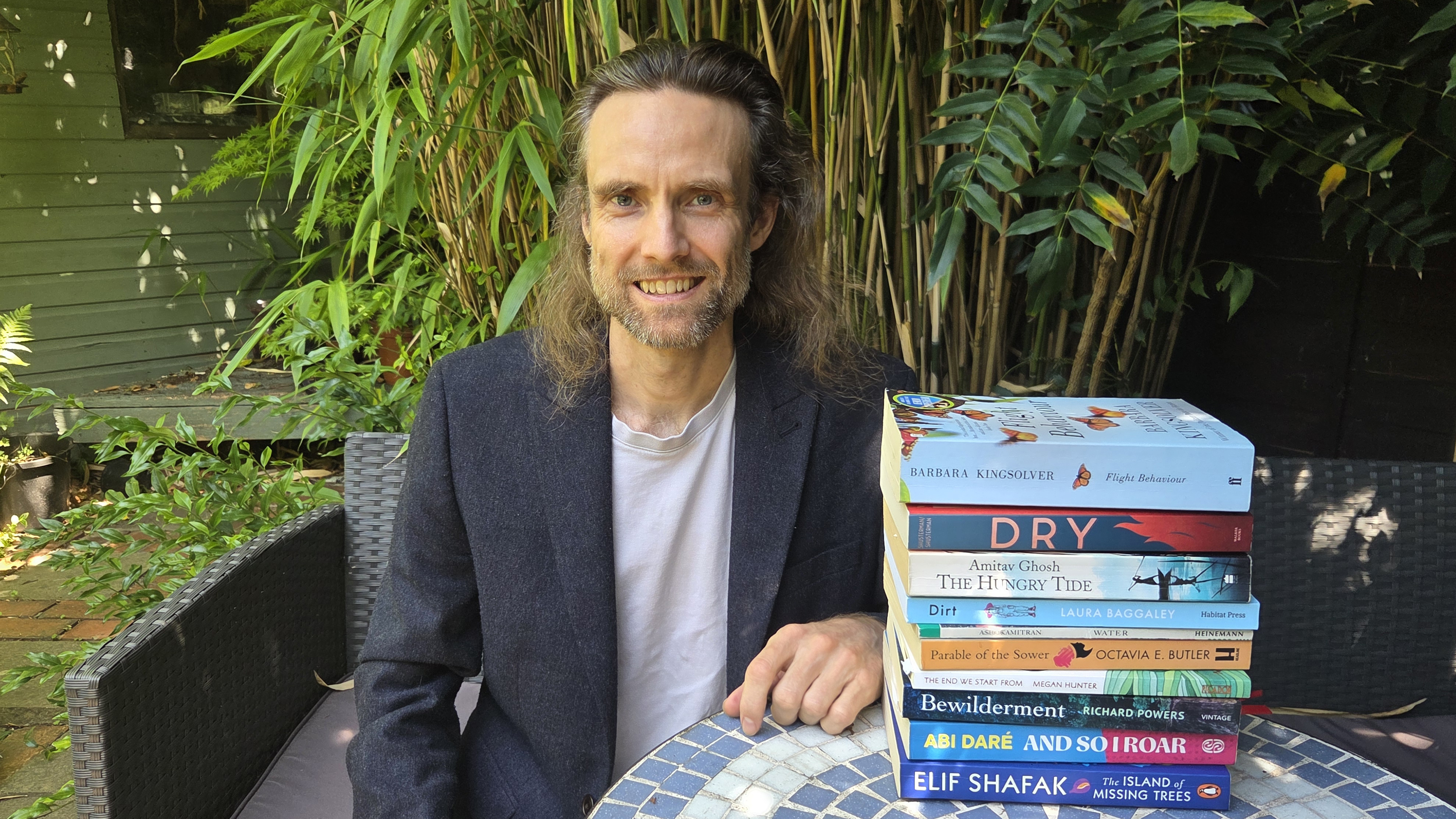 Dr Harvey is dressed in t shirt and dark suit jacket. He is outdoors seated on a chair next to a blue mosaic table. One the table are various climate fiction books stacked to make a tower.