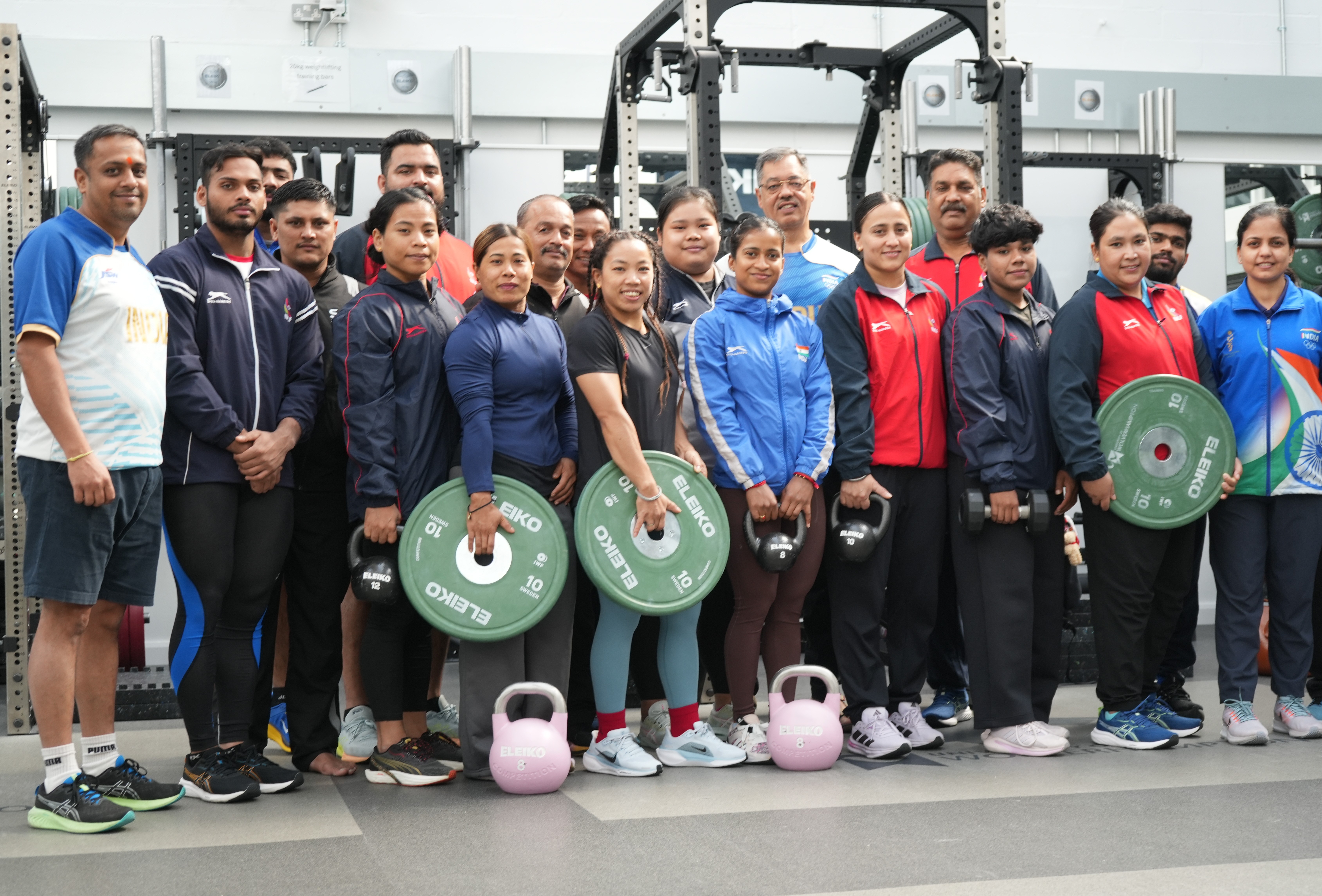 Eighteen athletes from the Indian Weightlifting Team line up for a group photo. They wear sports kit and are holding weights. Behind then are weightlifting machines.