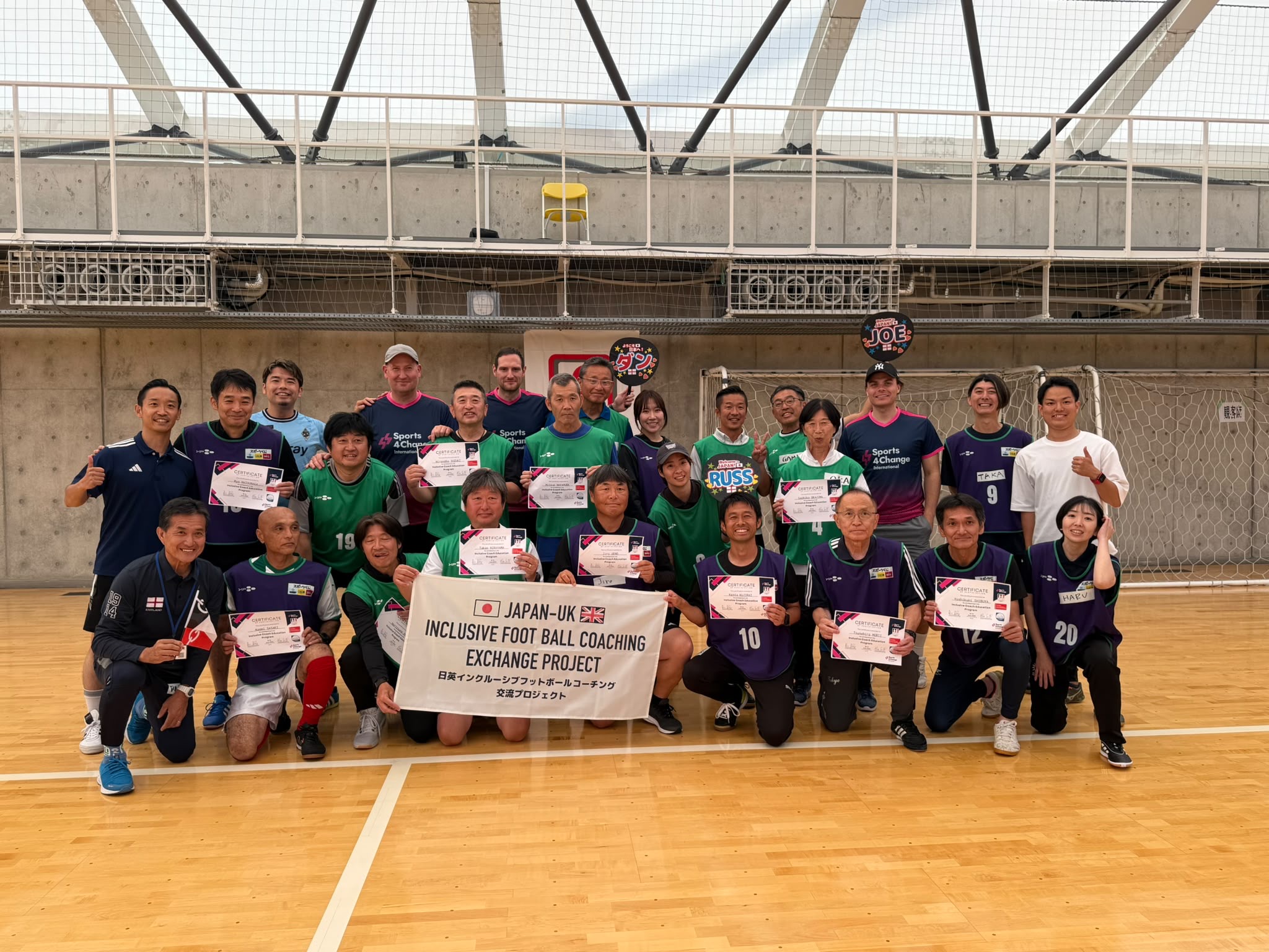 Japanese male footballers gathered in a sports hall for a group photo. They wear green football shirts and hold a banner that says 'Japan-UK Inclusive Football Exchange Project.'