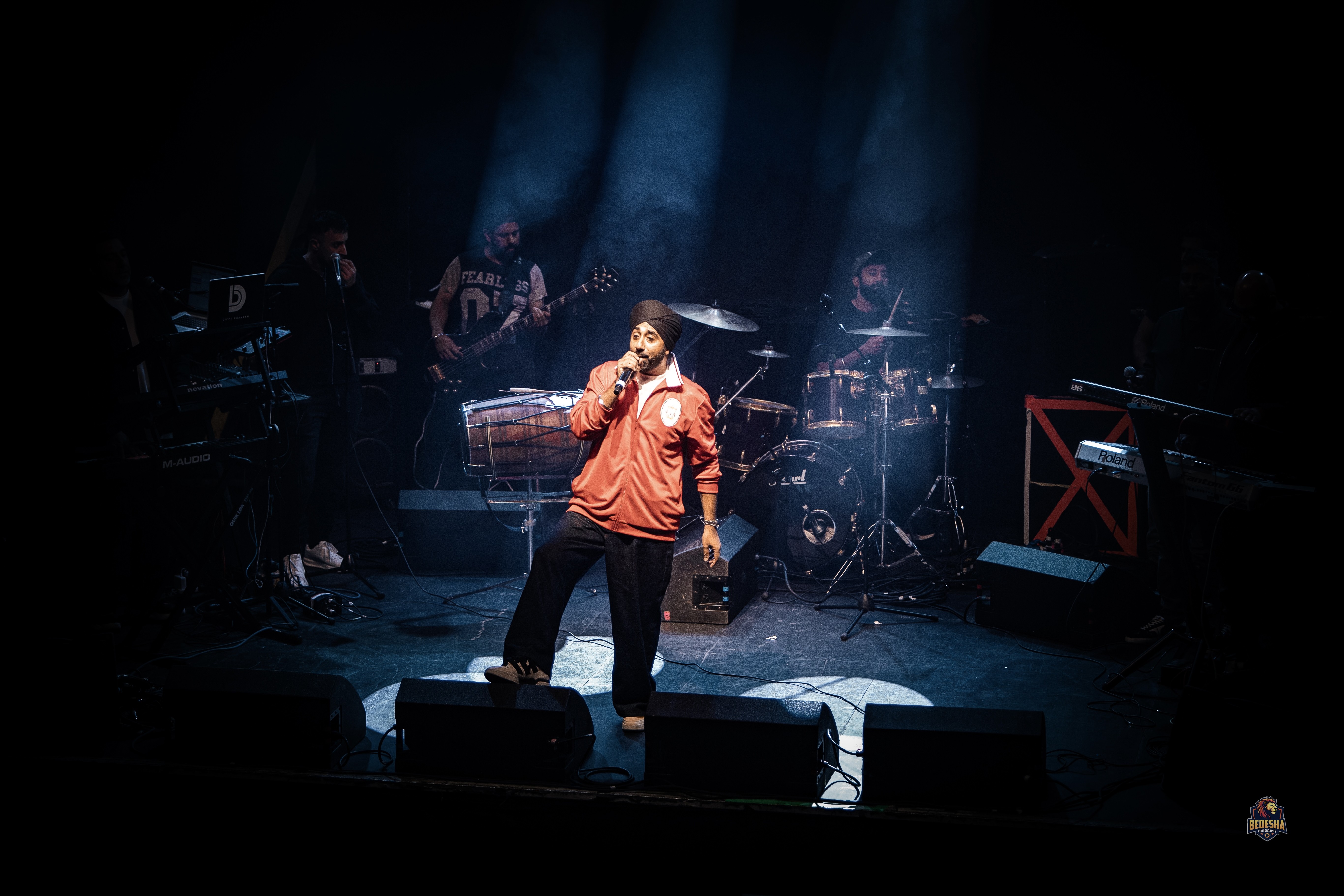 Jassi Sindhu pictured on-stage at the tribute event. He wears an orange jacket and black turban. He is in the spotlight, and the band can be seen around him in the shadows.