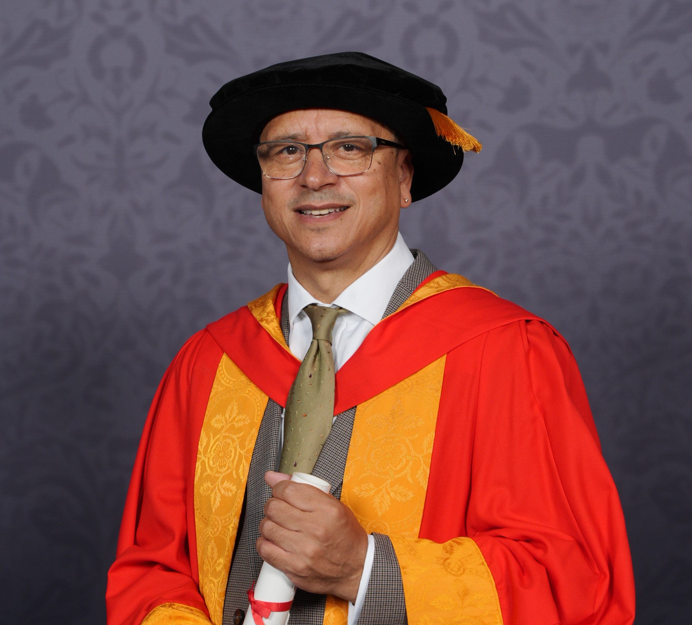 Jeremy poses for a professional graduation photo dressed in graduation robes in University of Wolverhampton colours, yellow and orange. He holds a scroll between his two hands and smiles for the camera.