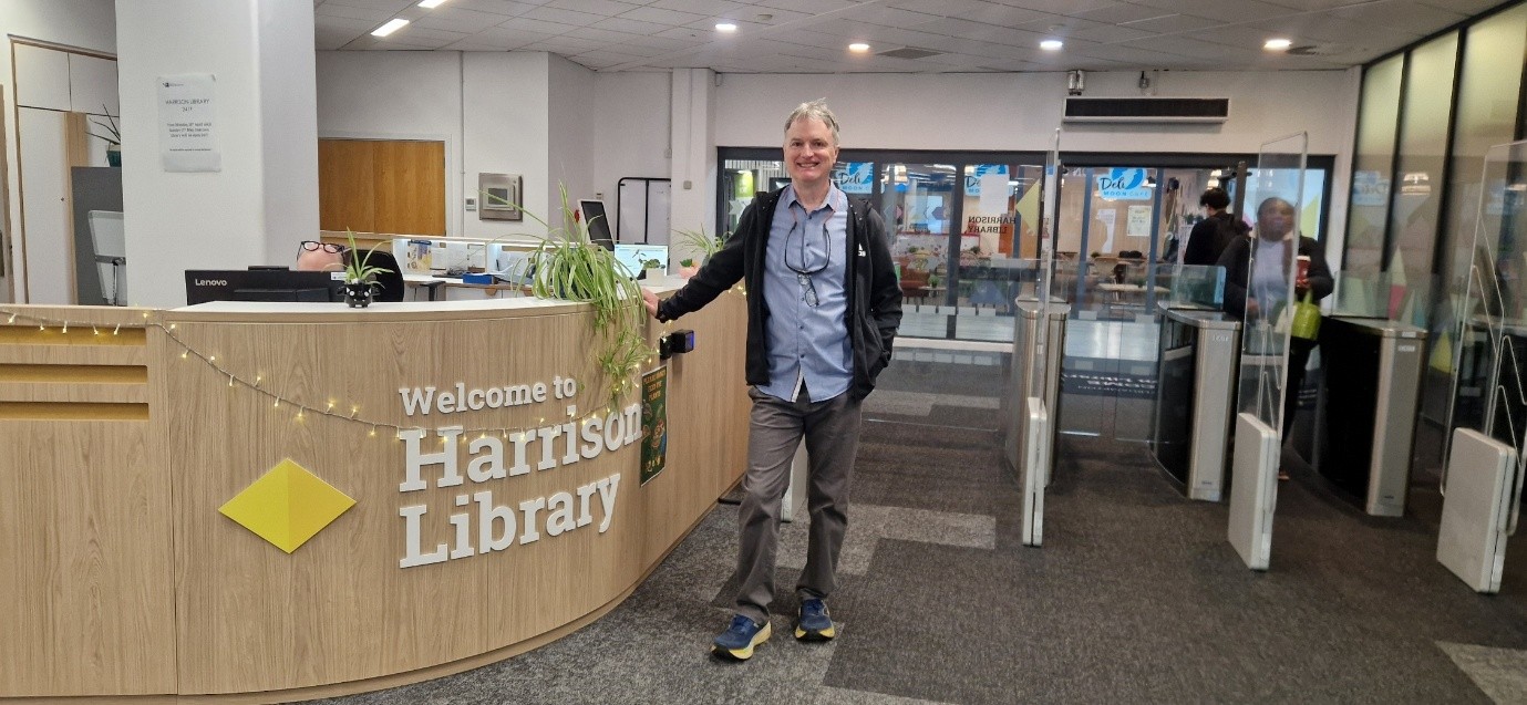 Joe stands at reception of Harrison Library at University of Wolverhampton. He smiles for the camera, wearing black jeans, blue shirt and dark blue casual jacket