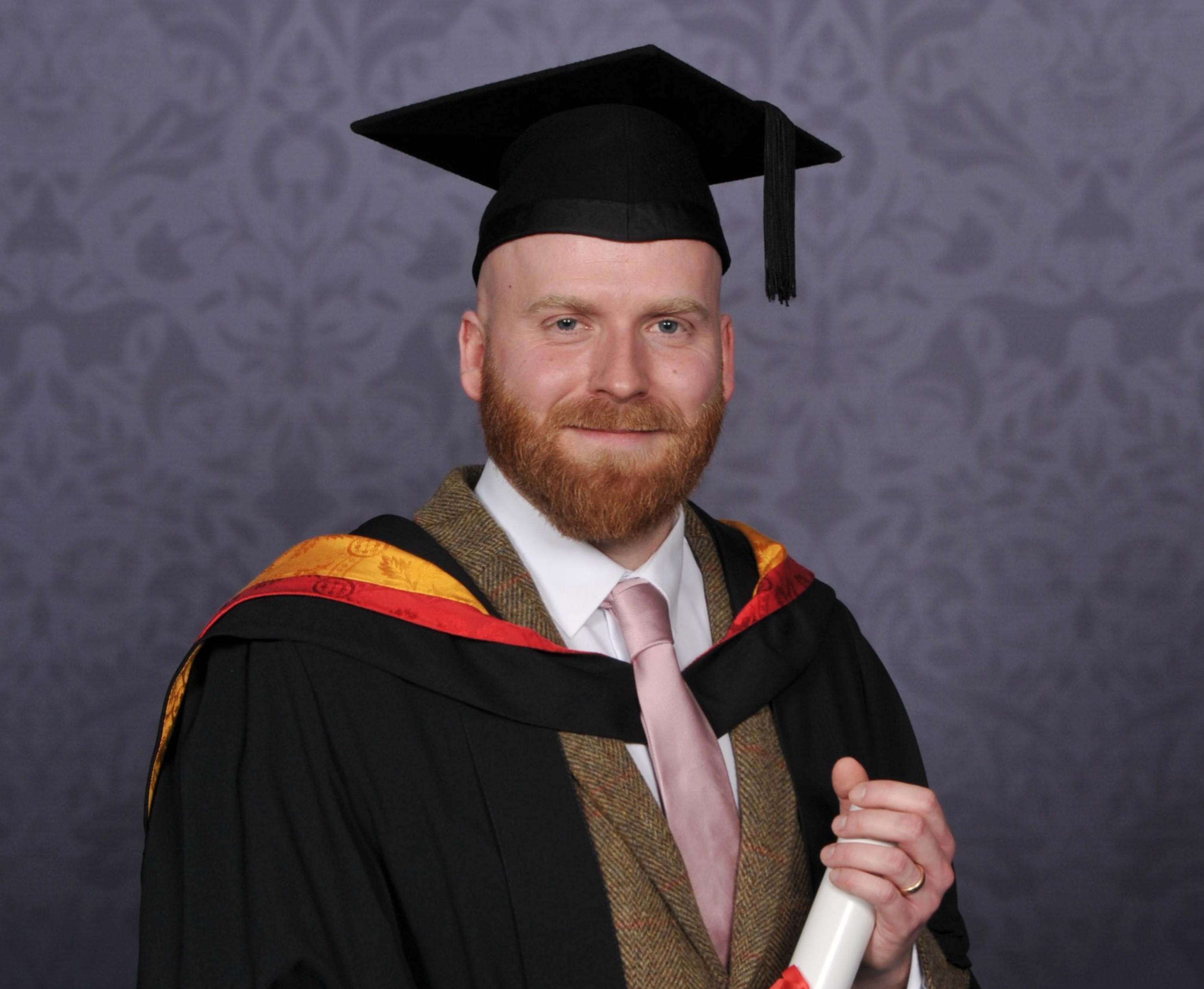 Joe poses for a professional graduation photograph in University of Wolverhampton orange and yellow gown. He holds a scroll between his hands.