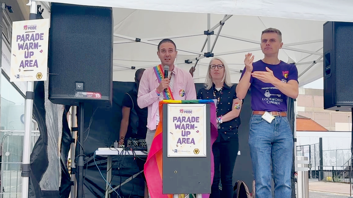 Josh Gulrajani stands next to Helen Keane on stage - on their right is a British Sign Language interpreter. Josh holds the mic and wears a pink shirt and Pride sash.