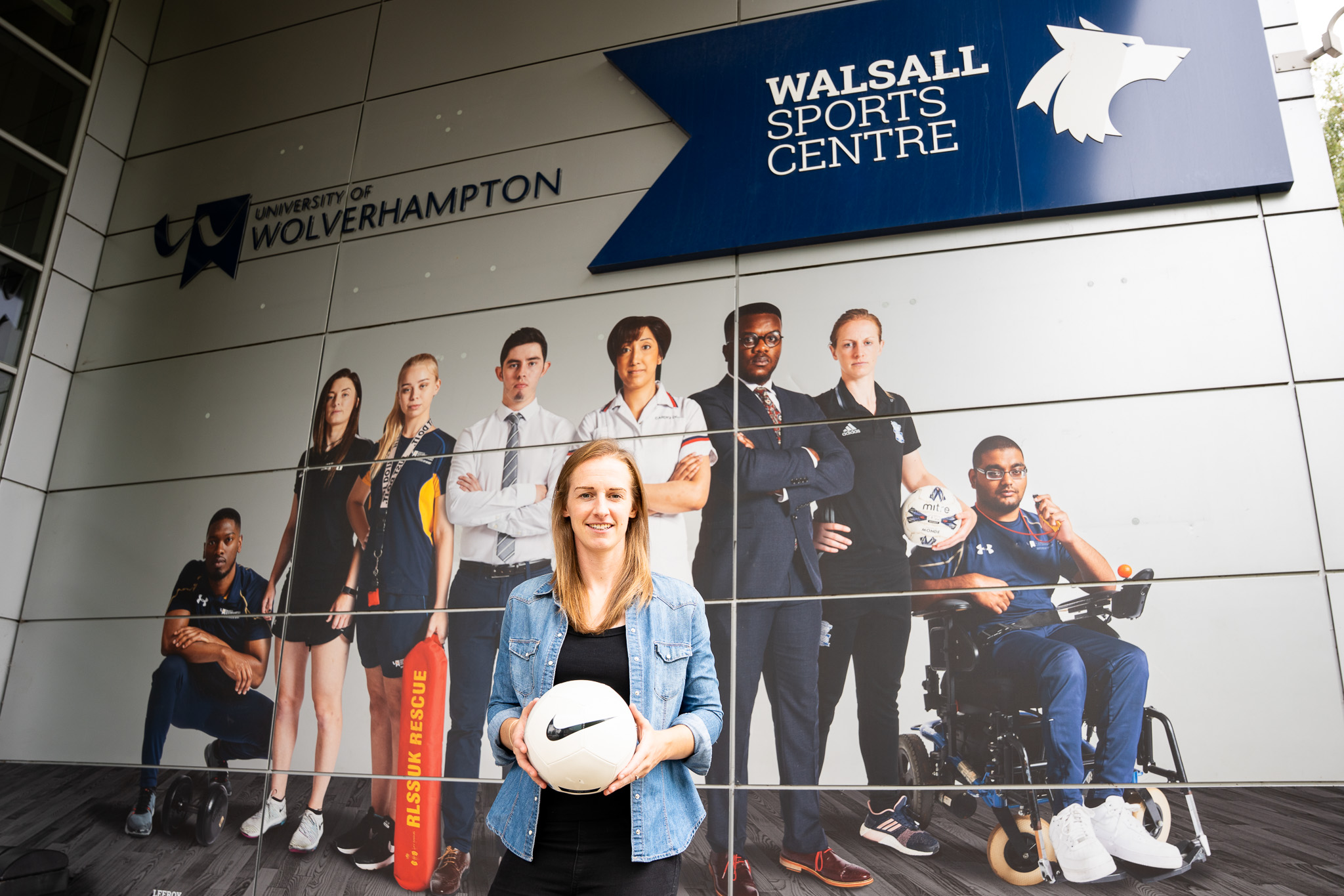 Kerys Harrop stands in front of at large photograph of sportspeople on a wall at Walsall Campus. She wears a denim shirt and is holding a Nike football.