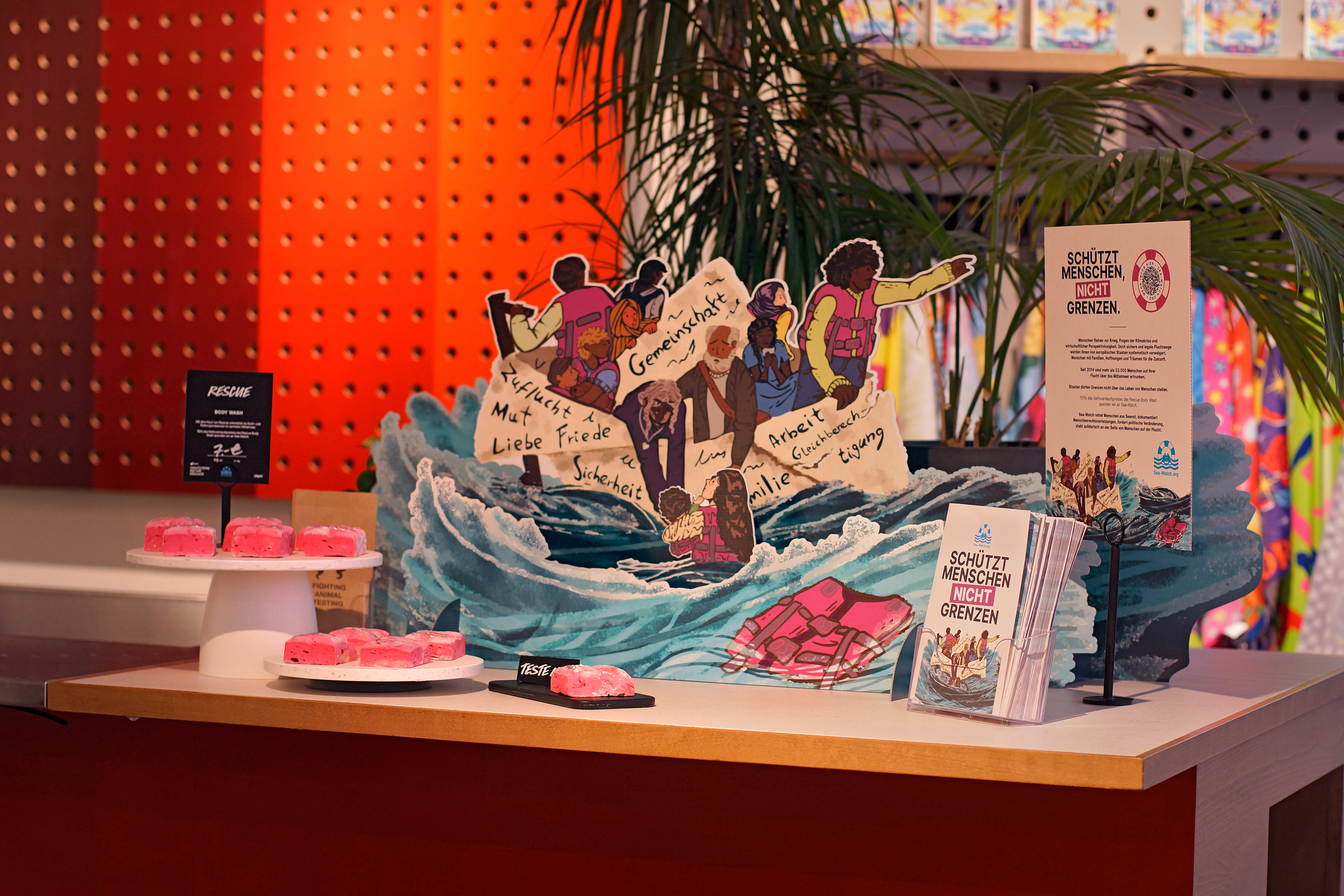 A display of marketing assets including leaflets and cardboard pop-ups are displayed on a table featuring Nayia's artwork. The pop up artwork is displayed as if on the sea.