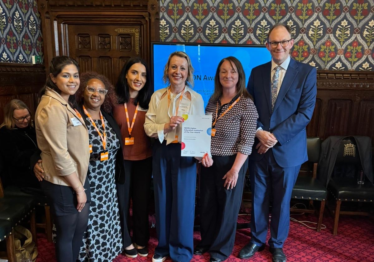 Five women and one man in a line at the NEON Awards wearing smart, casual attire. The woman in the centre holds a certificate and they all smile for the camera.