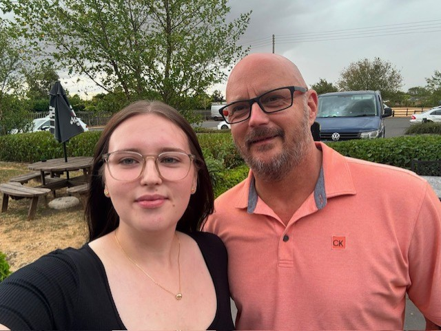 Chloe is in the foreground and her father, Peter stands slightly behind. They are outside, in front of a tree. Chloe wear a black top and Peter wears a salmon pink polo shirt and glasses