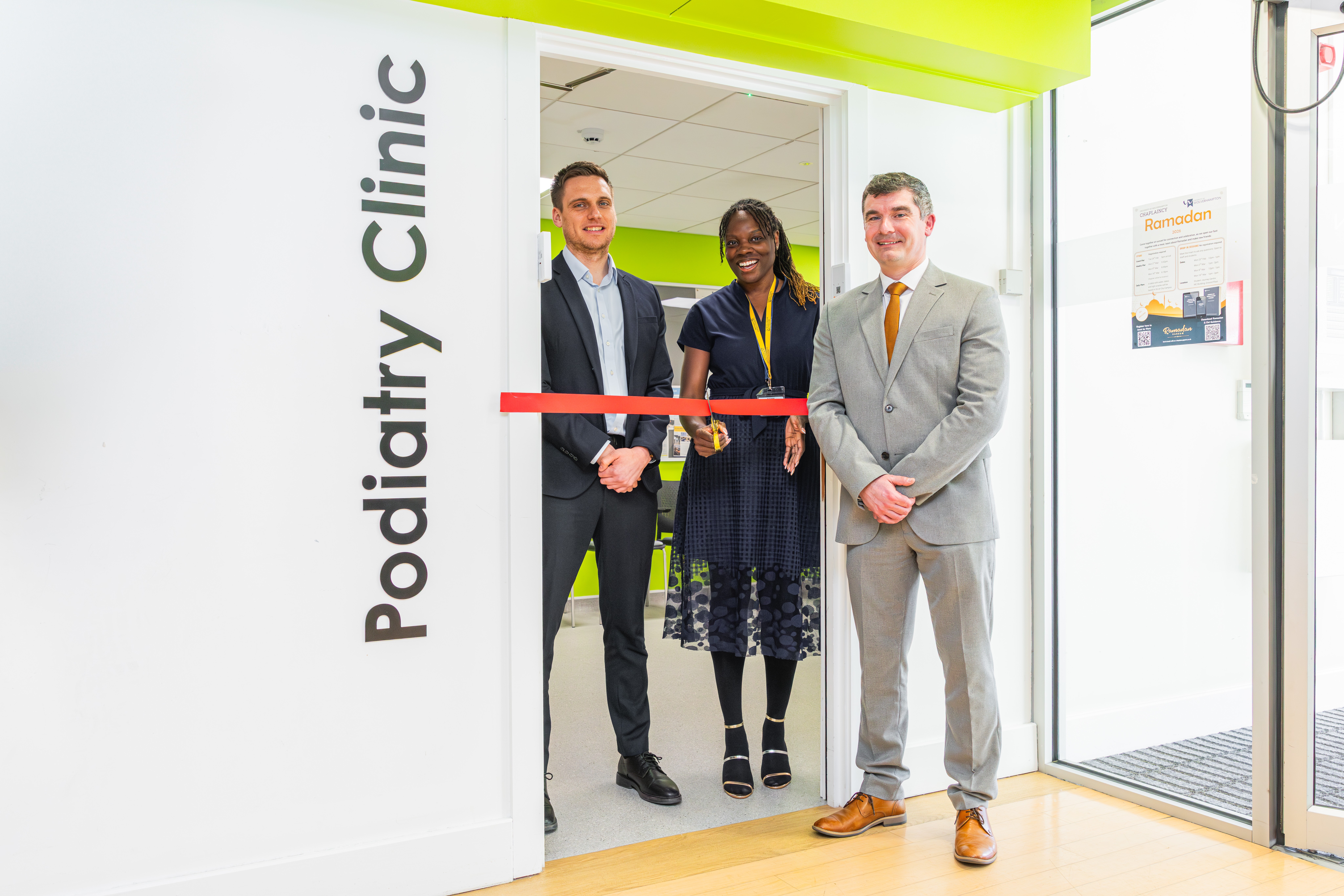 Two men dressed in suits flank a lady wearing a blue dress in the doorway of the new Podiatry Clinic. The open doorway is crossed with a red ribbon and the lady holds some scissors to cut the ribbon to officially open the facility.